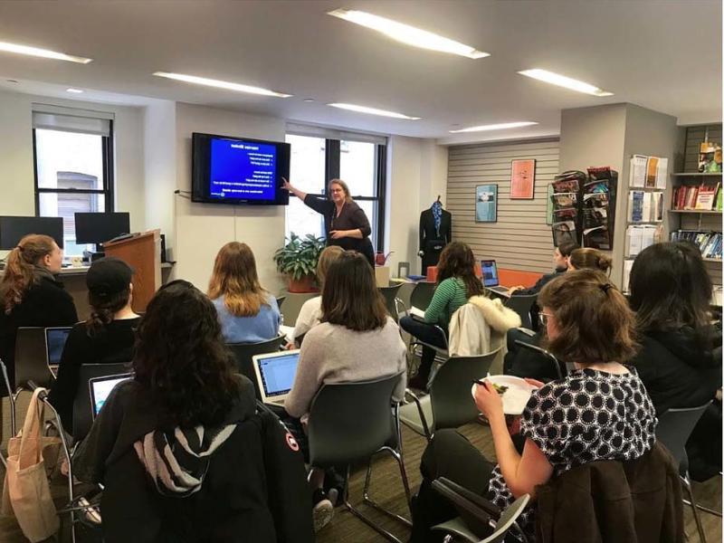 Woman pointing at screen while speaking to an audience of 12 or so students